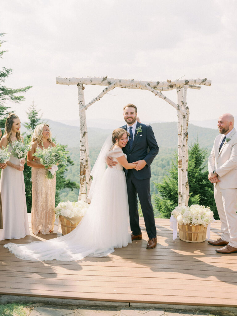 The bride wraps her arms around the groom in a joyful embrace under the birch arch at Oak Mountain, surrounded by bridesmaids holding bouquets.