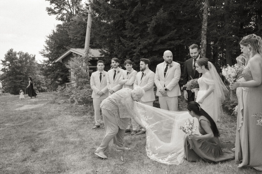 Black-and-white candid of bridesmaids and a family member adjusting the bride’s long veil while the groomsmen look on at Oak Mountain.
