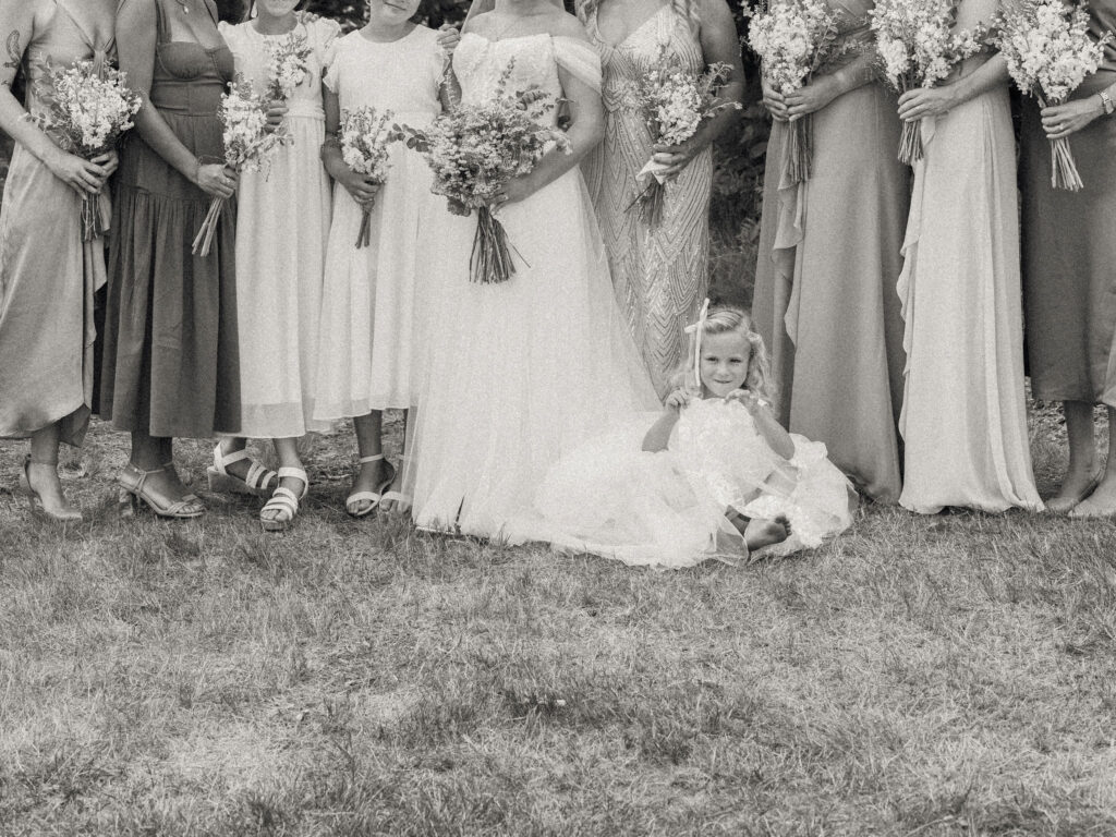 Black-and-white photo of a flower girl sitting barefoot in the grass in front of the bridal party, surrounded by bouquets and dresses.