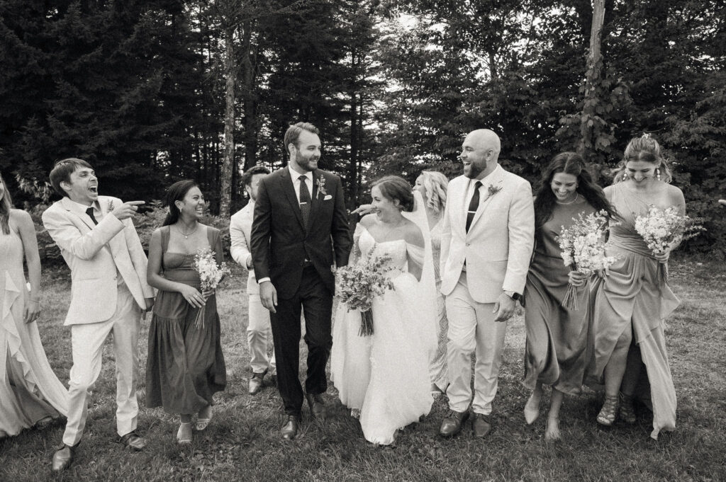 Black-and-white image of the full wedding party walking arm-in-arm through the grass, laughing and celebrating after the ceremony.
