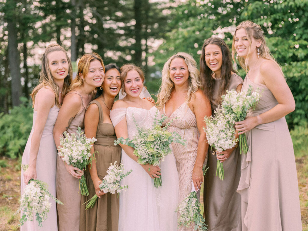 The bride stands smiling with her bridesmaids at Oak Mountain, all holding white-and-green bouquets against a backdrop of summer trees.