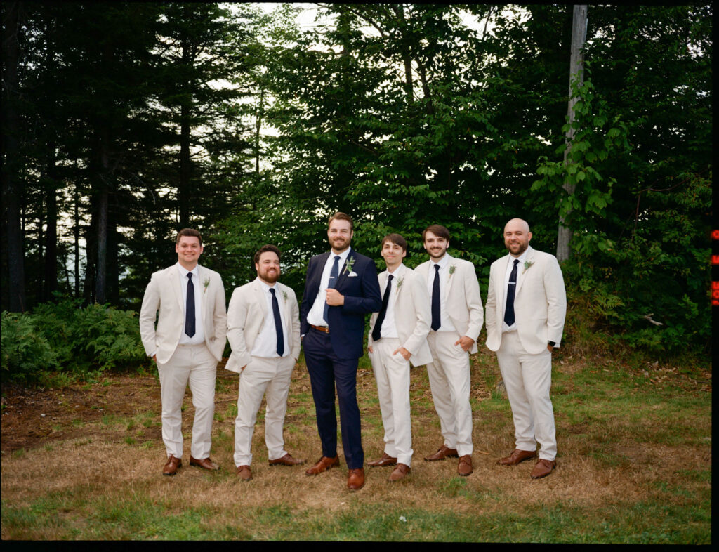 Film portrait of the groom and his groomsmen in light suits standing together at the forest edge, smiling toward the camera.