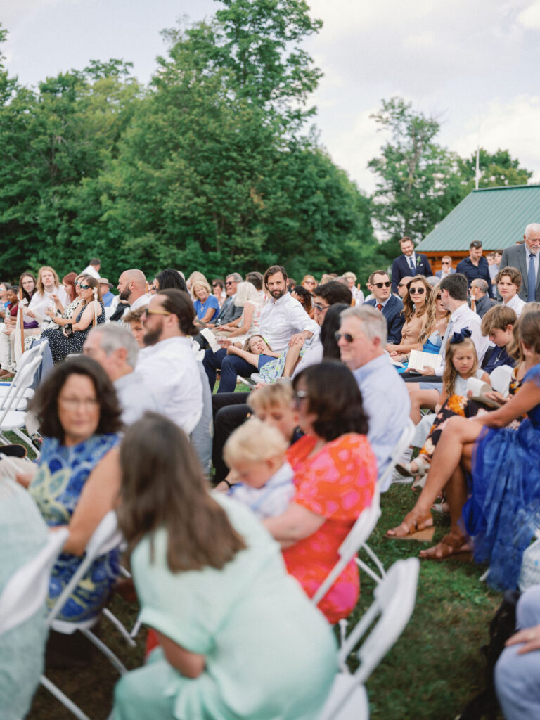 Wedding guests gather and chat in rows of white chairs before the ceremony begins, surrounded by summer greenery at Oak Mountain.