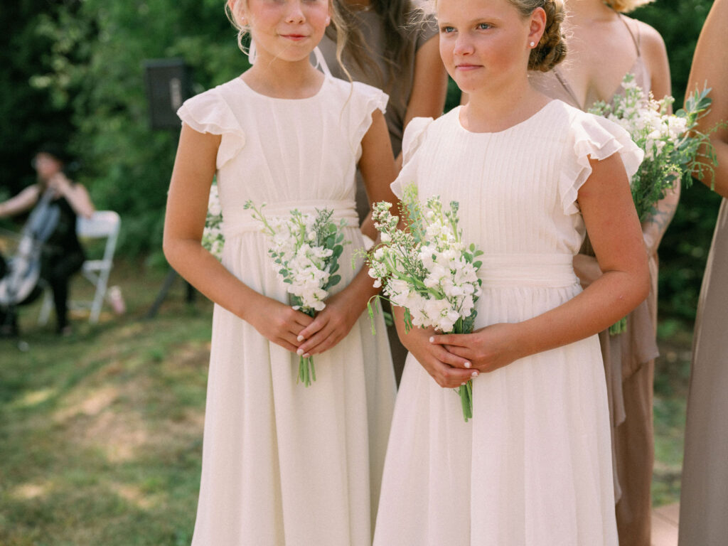 Two junior bridesmaids in cream dresses hold small white bouquets during the outdoor ceremony at Oak Mountain.