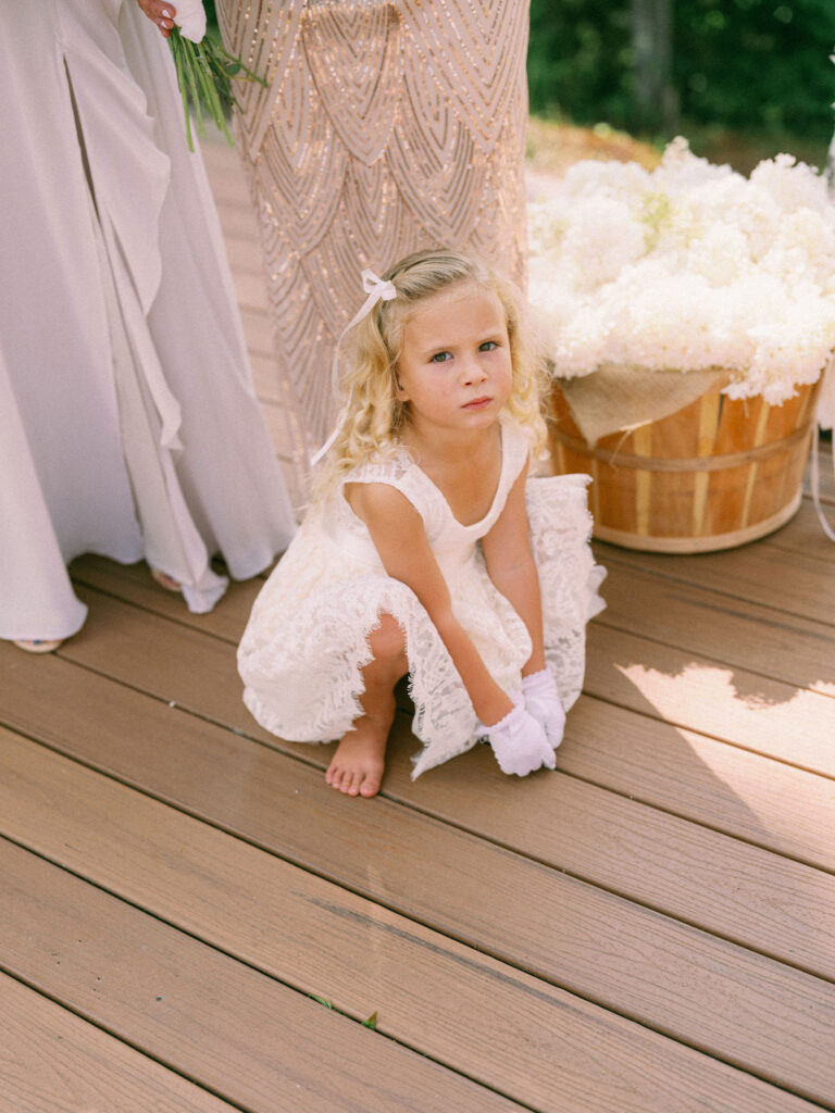 A barefoot flower girl in a lace dress crouches beside the bridesmaids on the ceremony platform, framed by a basket of white flowers.