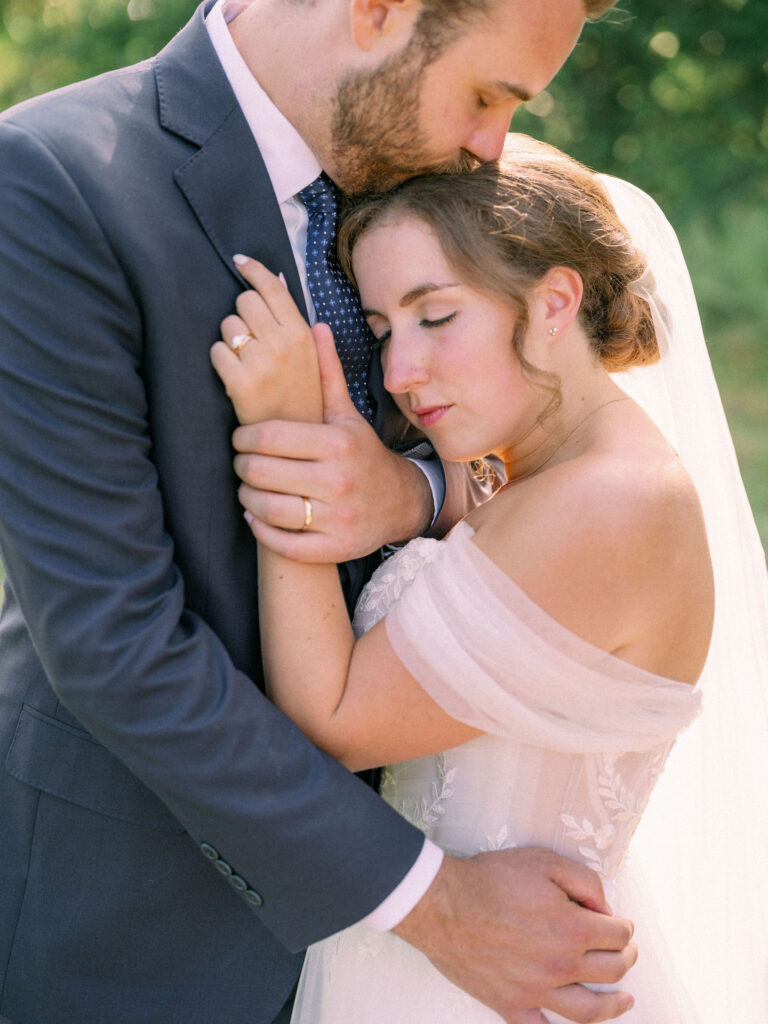 Bride and groom embracing closely outdoors, the groom kissing her forehead as she rests her eyes, holding his hands.