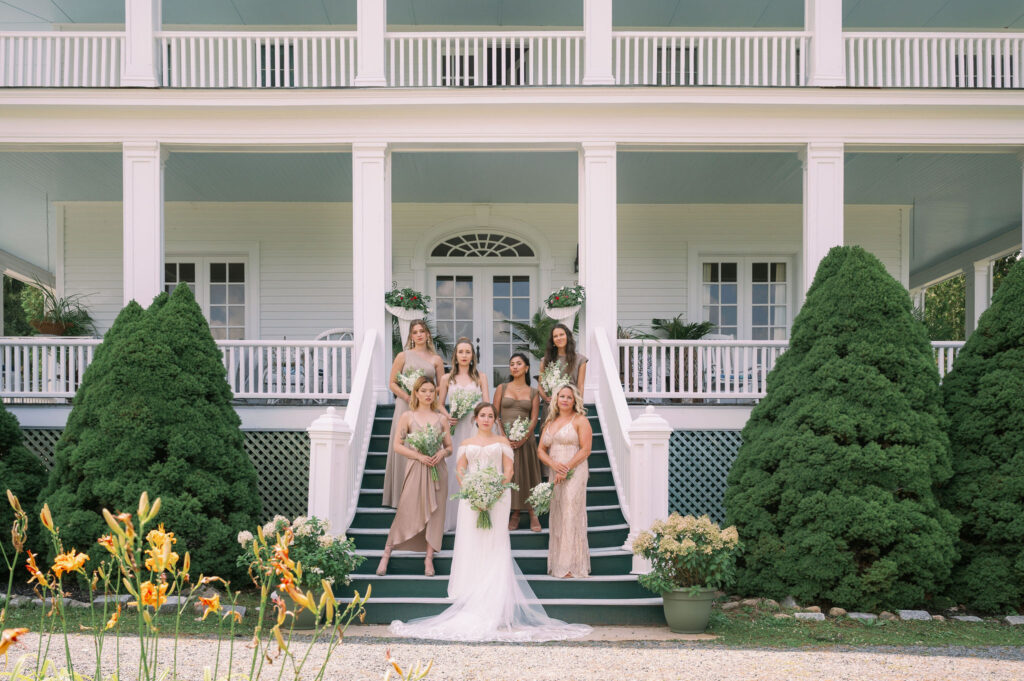 The bride and her bridesmaids stand on the front steps of The White House on the Lake, holding white-and-green bouquets against the estate’s grand white façade.