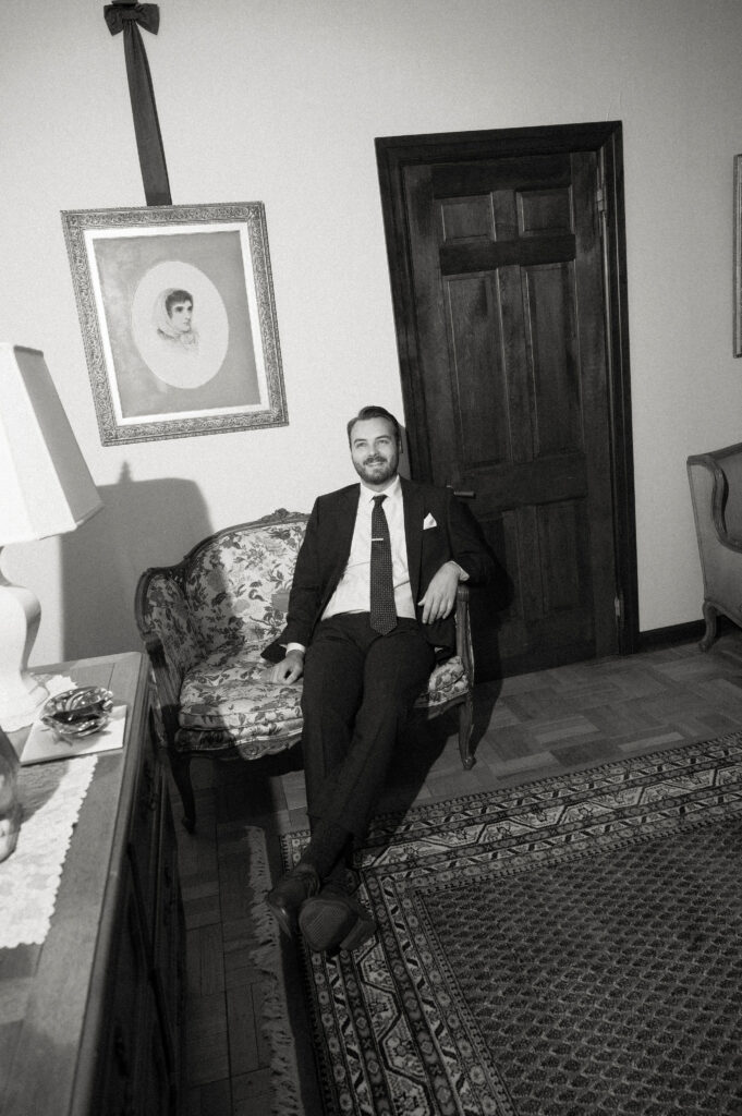 The groom relaxes on a floral loveseat in a vintage sitting room at The White House on the Lake, dressed in a suit and tie, surrounded by antique décor.