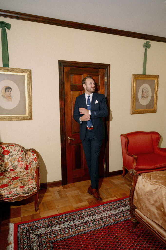 The groom stands in an elegant vintage bedroom at The White House on the Lake, adjusting his cuff while soft window light falls across the room.