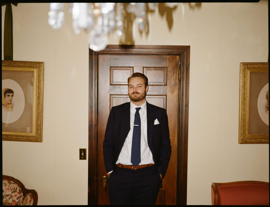 The groom stands in a vintage room at The White House on the Lake, wearing a navy suit and tie, framed by wood paneling and antique portraits.