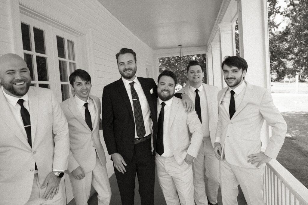 The groom and his groomsmen laugh together on the porch of The White House on the Lake, dressed in coordinating suits during the wedding morning.
