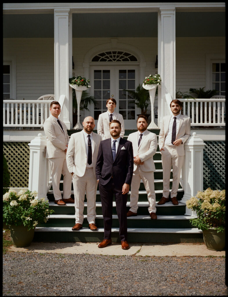 The groom and his groomsmen pose on the front steps of The White House on the Lake, dressed in classic suits against the historic white estate façade.