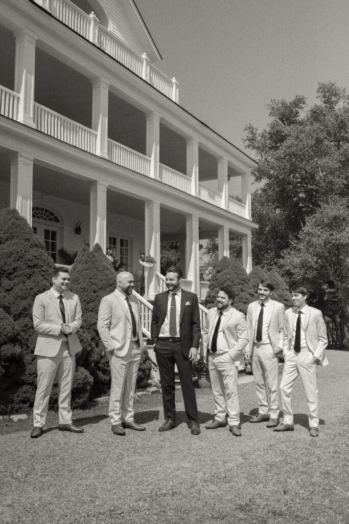 The groom and groomsmen stand together in front of the grand white façade of The White House on the Lake, dressed in light and dark suits under bright summer light.