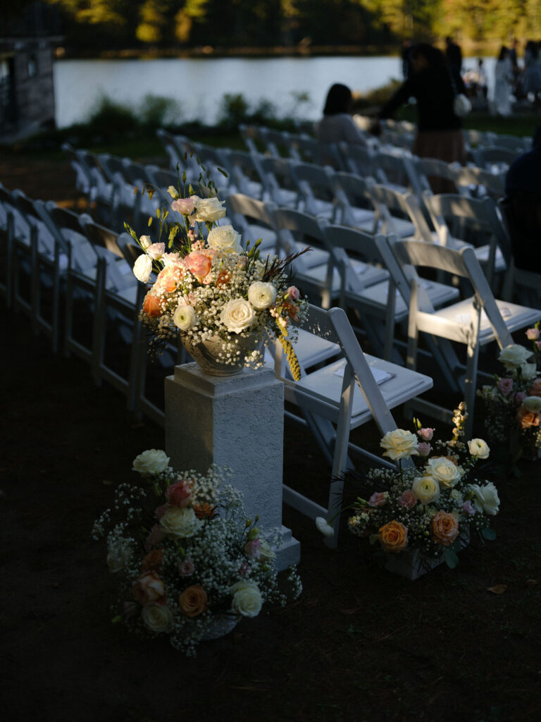 Floral arrangements line the aisle at an outdoor lakeside wedding ceremony in Lake Placid, with white chairs arranged facing the water.