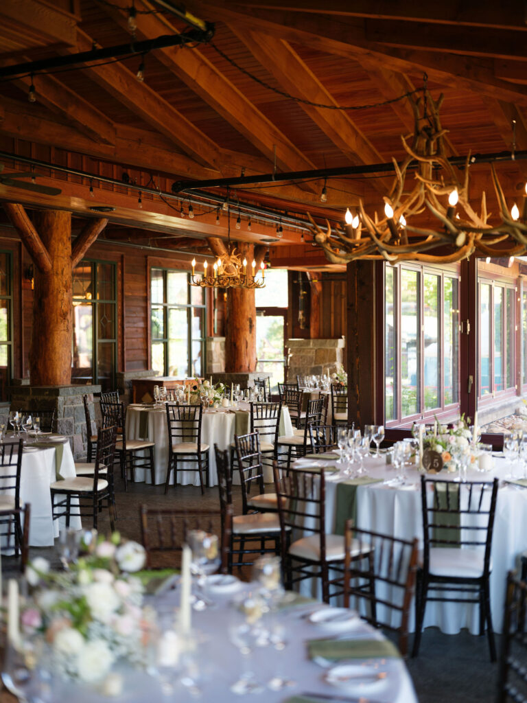 Wedding reception space at the Whiteface Lodge featuring round tables, antler chandeliers, and warm wood beams in a rustic Adirondack setting.