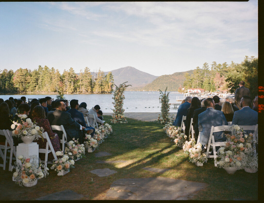 Outdoor wedding ceremony at Canoe Club set along the lakeshore in Lake Placid, with floral-lined aisle, seated guests, and Adirondack mountains in the background.