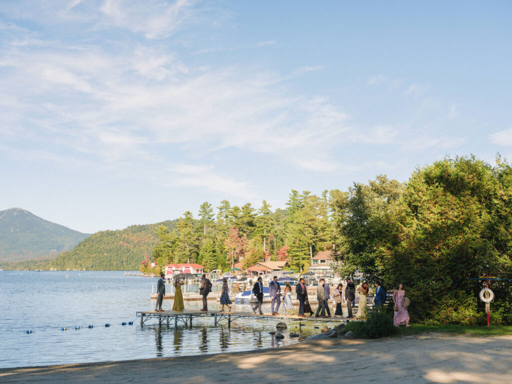 Guests walk along a wooden dock beside the lake during a fall wedding in Lake Placid, with boats, trees, and mountains surrounding the shoreline.