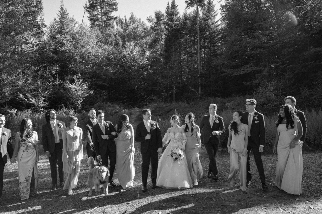 Wedding party walks together through a forest clearing during fall wedding portraits, captured in black and white with dappled sunlight.