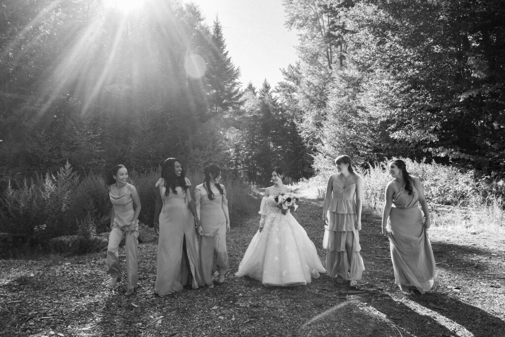 Bride walks alongside her bridesmaids through a forest clearing during fall wedding portraits, captured in black and white with sun filtering through the trees.