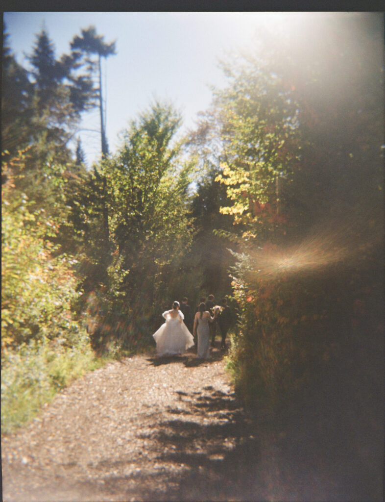 Bride follows behind the wedding party along a wooded trail during fall wedding portraits, captured on film with soft light and autumn foliage.