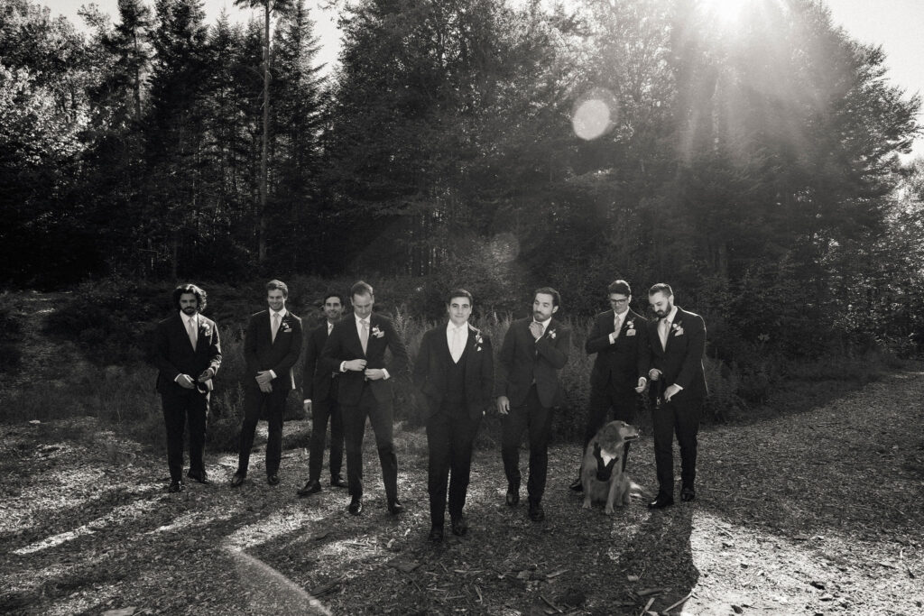 Groomsmen gather together during fall wedding portraits with the couple’s dog dressed in a vest, captured in black and white outdoors.