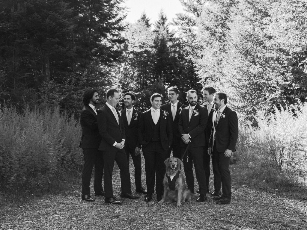 Groomsmen stand together with the couple’s dog during fall wedding portraits, captured in black and white in a forest clearing.