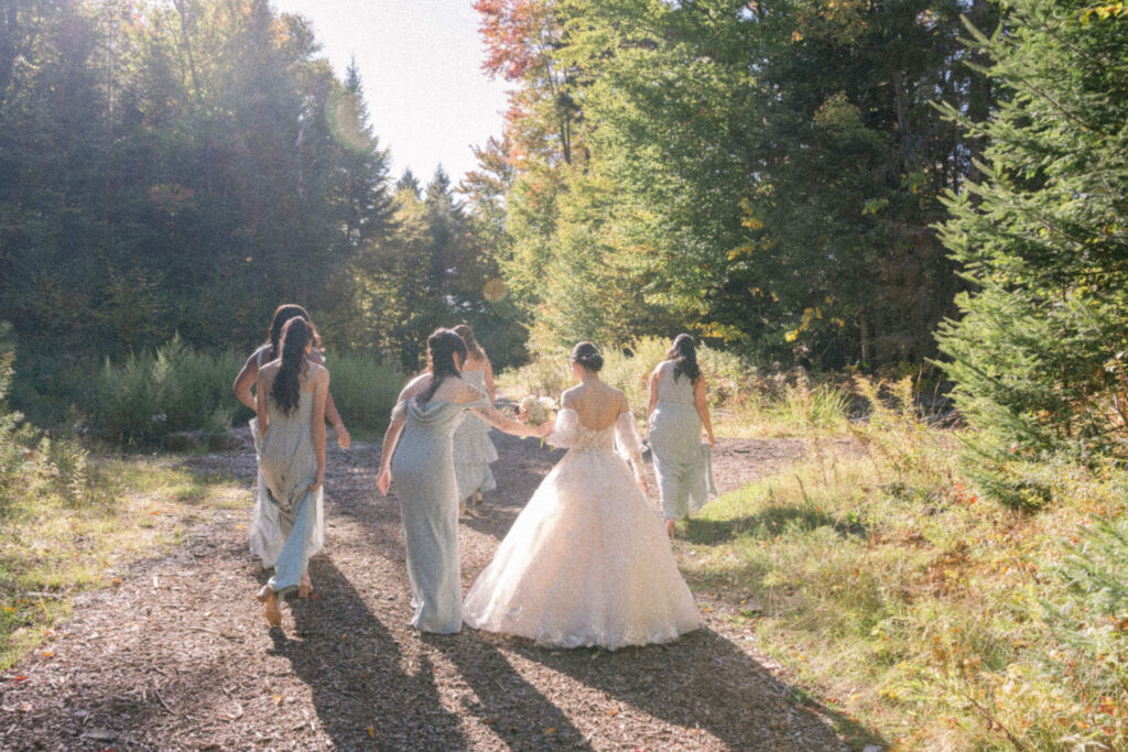 Bride walks hand in hand with her bridesmaids along a wooded path during fall wedding portraits, captured on film in soft afternoon light.