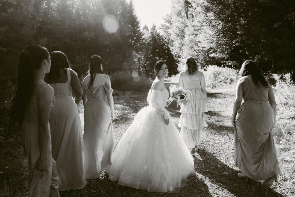 Bride glances back toward the camera while walking with her bridesmaids along a wooded trail, captured in black and white during fall wedding portraits.