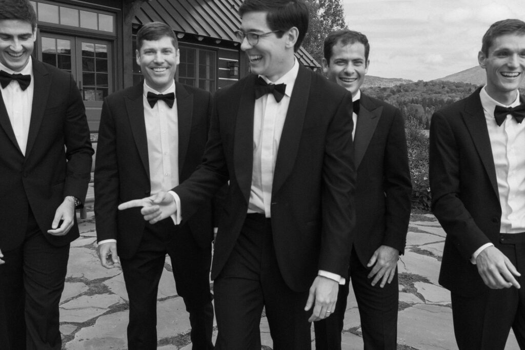 Groomsmen in black tuxedos walk and laugh together outside a rustic Lake Placid, New York wedding estate with Adirondack mountain views.