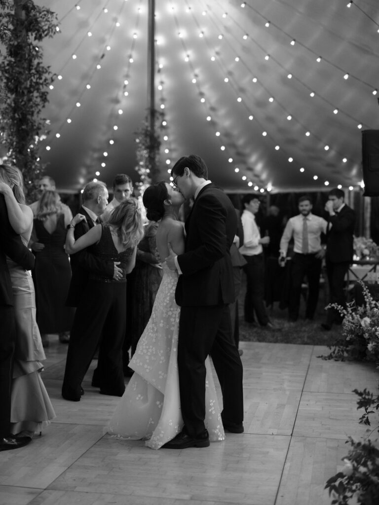 Bride and groom share a kiss on the dance floor beneath string lights inside a wedding tent in Lake Placid.