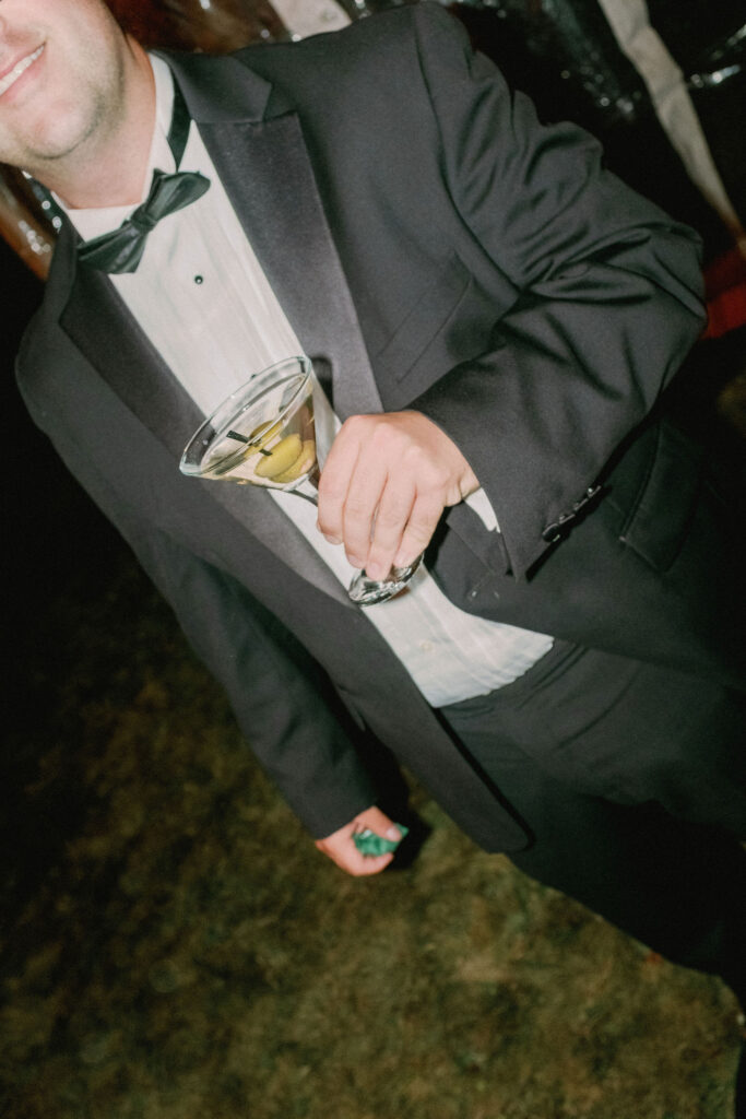 Wedding guest in a tuxedo holds a martini while walking outside during a Lake Placid wedding reception.