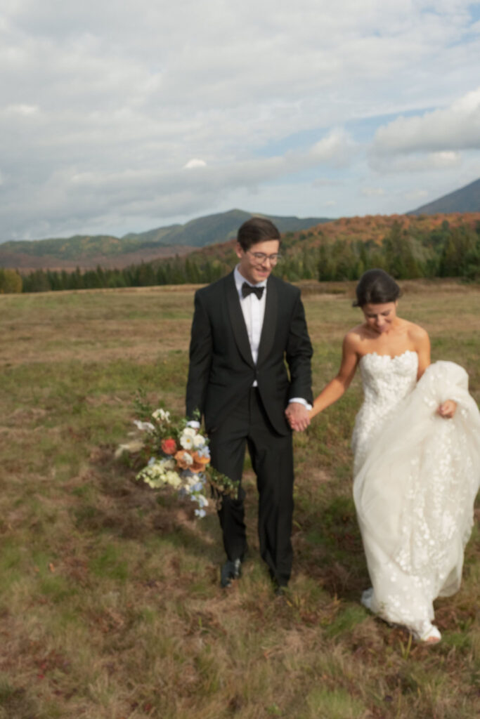 Bride and groom walk hand in hand through a grassy Adirondack field during their Lake Placid wedding portraits.