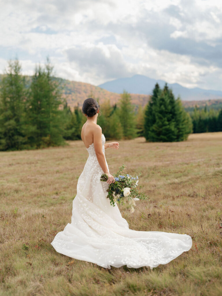 Bride in a flowing lace gown holds a bouquet while looking toward Adirondack mountains during a Lake Placid wedding.