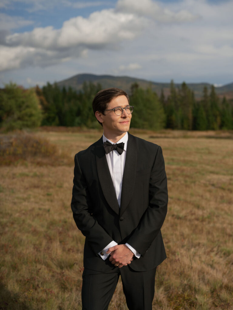 Groom in a black tuxedo stands in an open Adirondack field with Lake Placid mountain views in the background