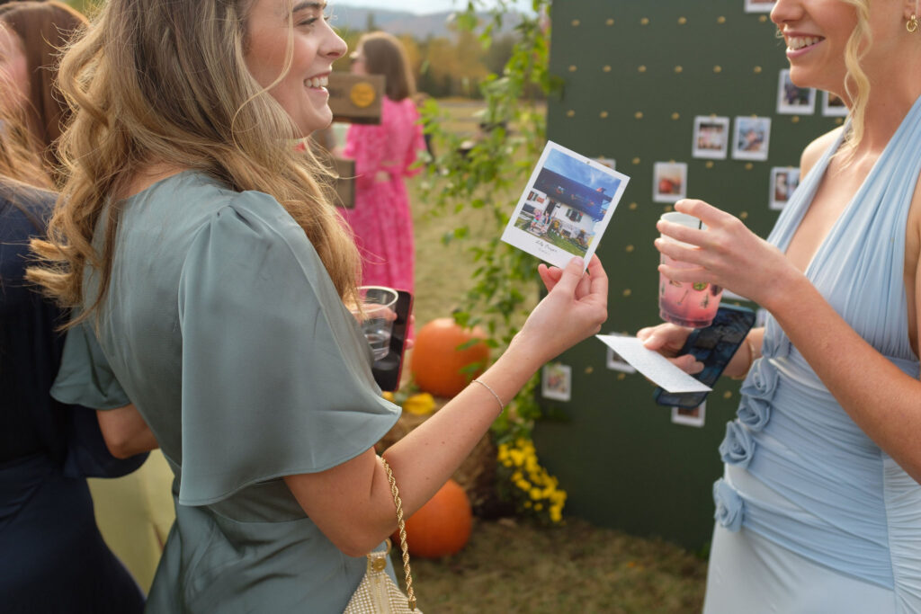 Wedding guests laugh while looking at Polaroid photos on a display board during cocktail hour at a Lake Placid, New York wedding.