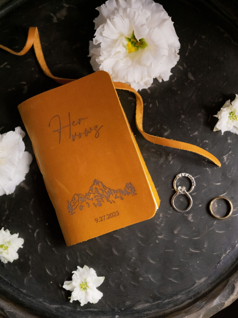 Lake Placid private estate wedding details with vow book, rings, and floral accents on dark tray