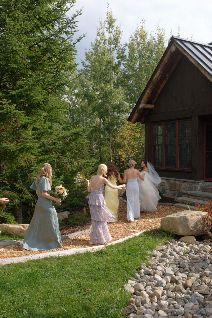 Bride and bridesmaids walk along a garden path beside a rustic estate in Lake Placid, New York, surrounded by trees and natural landscape.