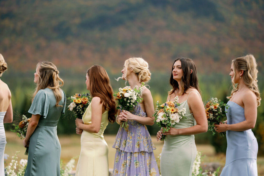 Bridesmaids in soft, mismatched dresses hold bouquets and stand in line during a mountain-view wedding ceremony at a private Lake Placid estate in the Adirondacks.