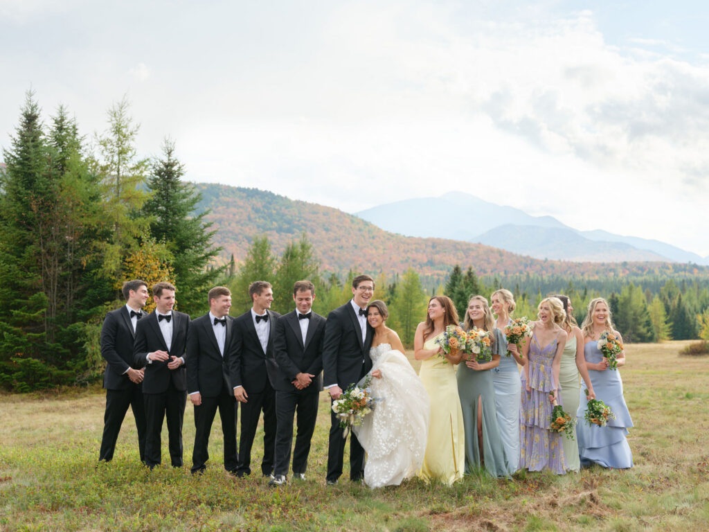 Bride, groom, bridesmaids, and groomsmen walk together across a field with Adirondack mountains during a Lake Placid wedding.