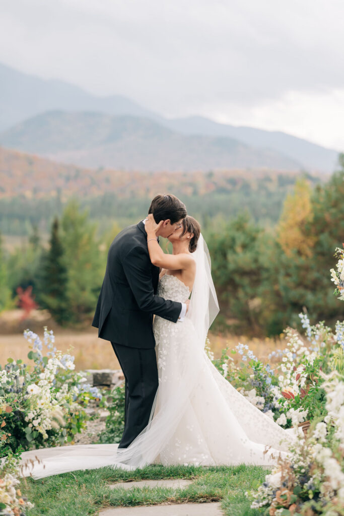 Bride and groom share their first kiss during an outdoor ceremony at a private estate in Lake Placid, New York, framed by garden florals and mountain views.