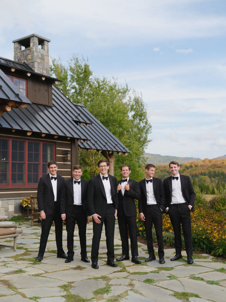 Groom and groomsmen pose in black tuxedos outside a rustic private estate in Lake Placid, New York, with Adirondack mountains in the background.