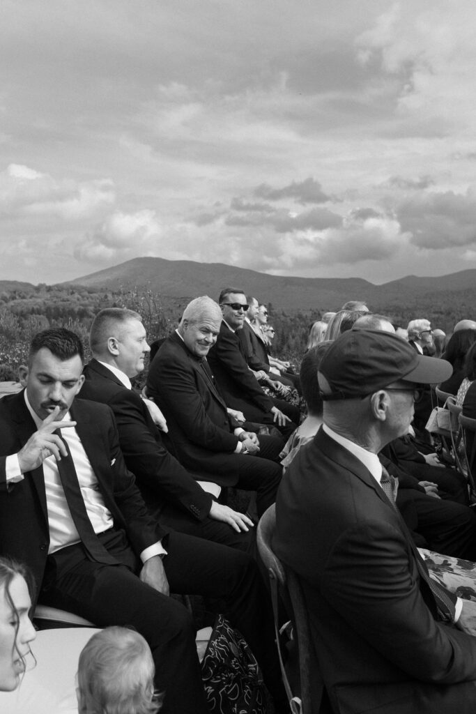 Wedding guests seated outdoors during a Lake Placid ceremony, captured candidly in black and white with Adirondack mountains in the background.