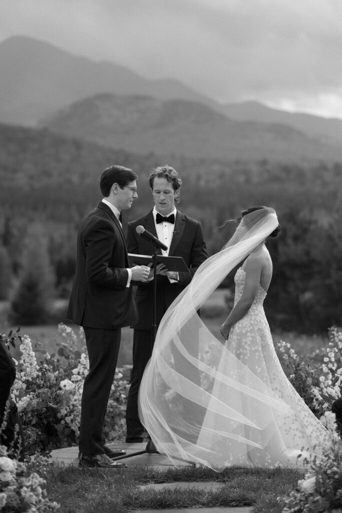 Black and white image of a bride and groom exchanging vows as the bride’s veil moves in the wind during a mountain-view ceremony in Lake Placid.