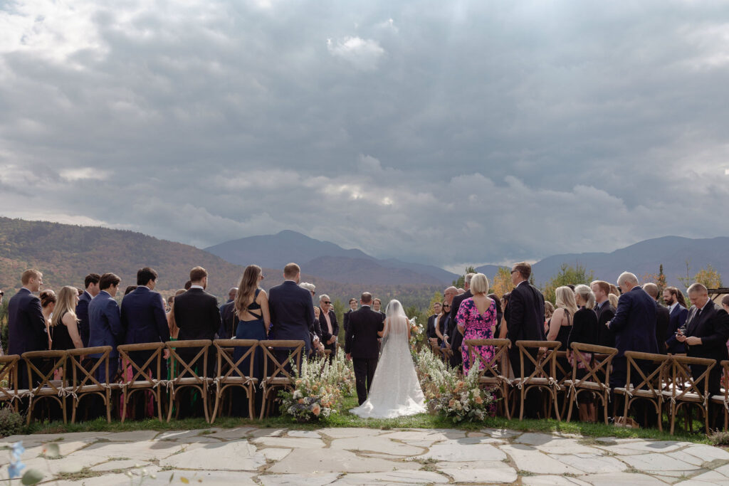 Wide view of an outdoor wedding ceremony aisle at a private Lake Placid estate, with guests standing and Adirondack mountains under dramatic skies.