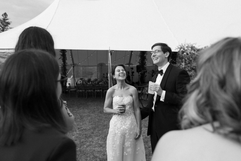 Bride and groom laugh together while chatting with guests outside a sailcloth tent at a Lake Placid wedding.