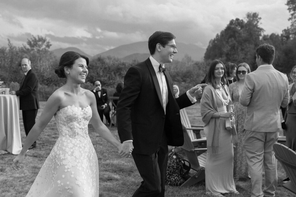 Bride and groom hold hands and walk through cocktail hour guests at an outdoor Lake Placid wedding.