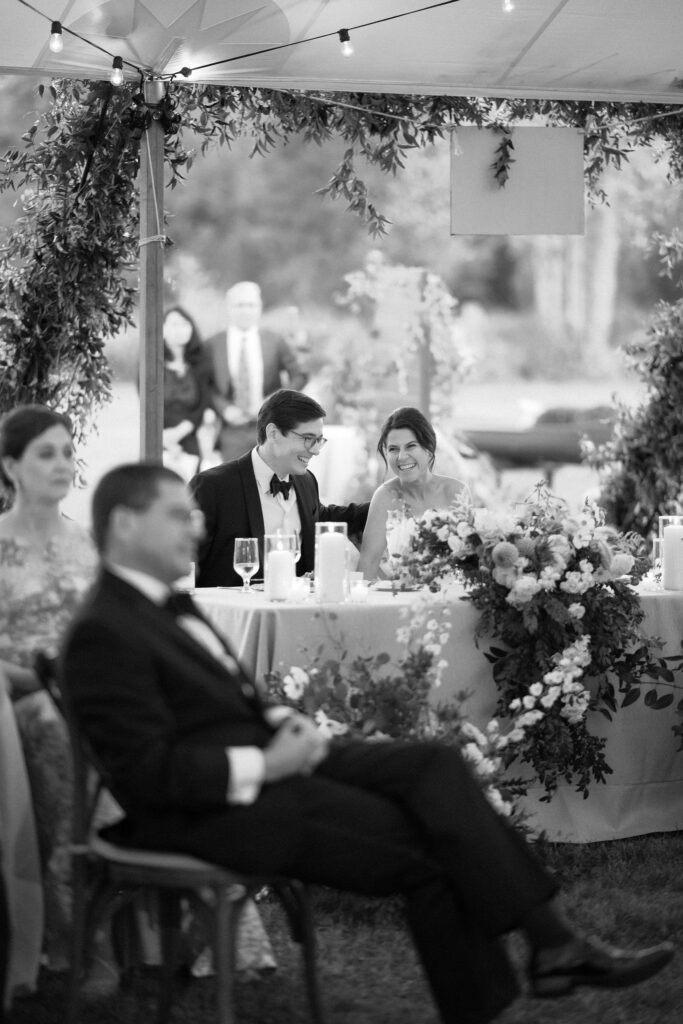 Bride and groom smile and laugh at their sweetheart table during a tented wedding reception in Lake Placid, New York.