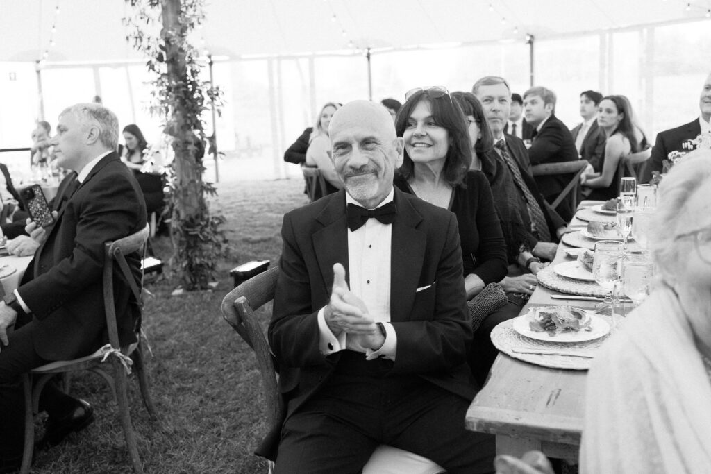 Wedding guest in a tuxedo claps during dinner speeches inside a sailcloth tent at a Lake Placid wedding.