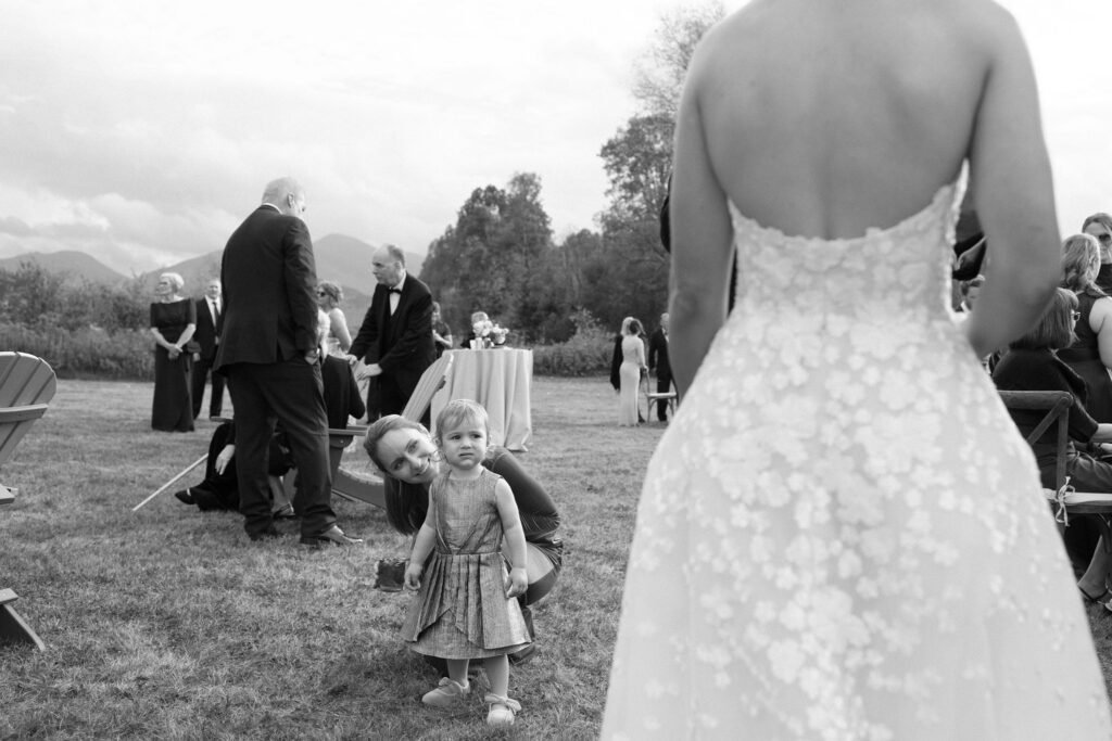 Bride in a lace gown walks past a small child and guests during an outdoor wedding in Lake Placid, New York.