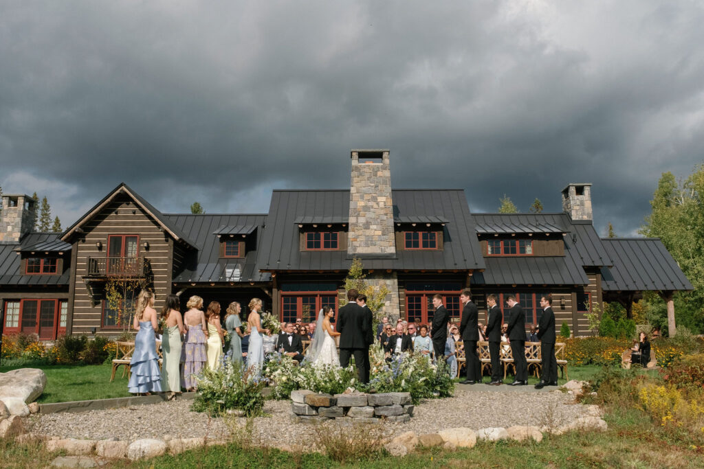 Outdoor wedding ceremony at a private estate in Lake Placid, New York, with a large rustic home, garden florals, and Adirondack mountains in the background.